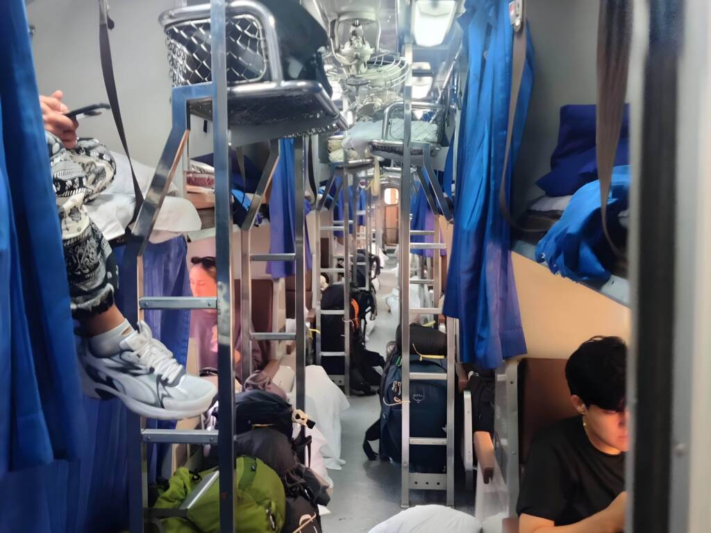 Interior of a crowded, second-class overnight sleeper train in Thailand, showing backpackers using cheap transport for budget travel.