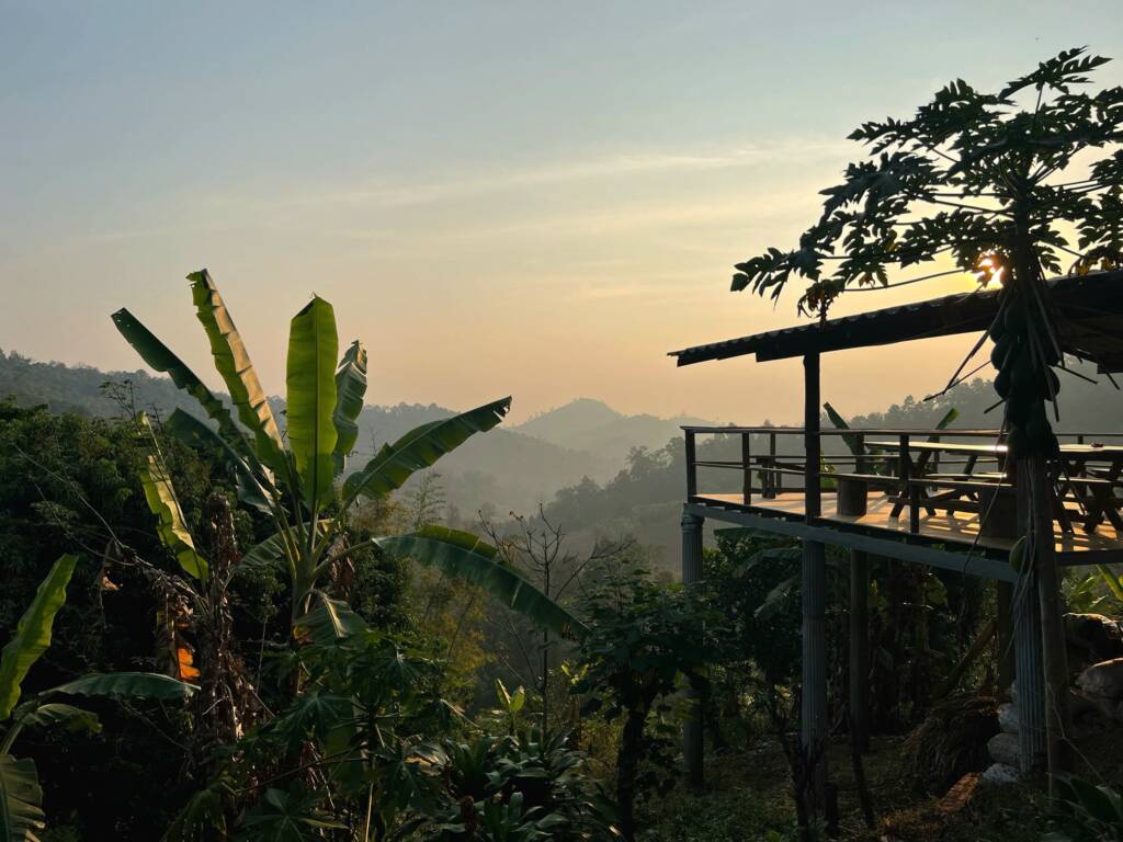 A tranquil sunset view from a jungle homestay balcony over the misty hills of Northern Thailand, highlighting the slow travel experience.
